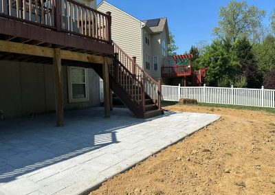 Backyard with a new stone patio, dirt border, wooden deck, white fence, and playset in the background.