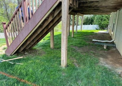 Wooden stairs and deck with grass and dirt underneath, next to a house with a white fence in the background.