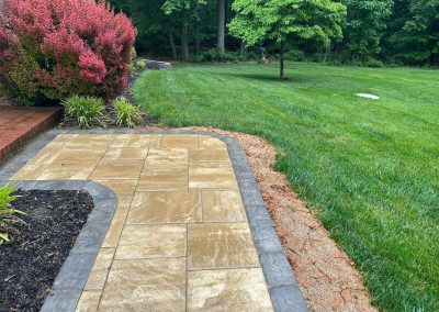 Wet stone patio with bordered edge beside green grass lawn, red bushes, and a tree in a backyard.