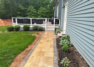 A stone walkway beside a blue house leads to a white-railed deck, with plants along the edge.
