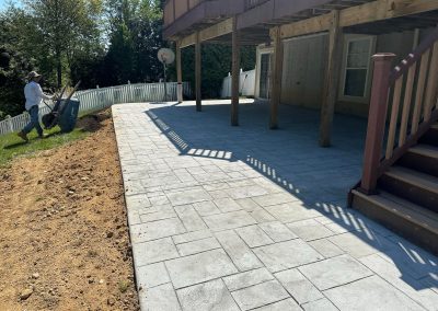 A man with a wheelbarrow works near a newly paved patio under a wooden deck on a sunny day.