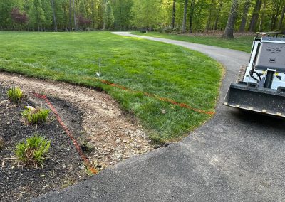 A marked section of grass by a driveway, with construction equipment and flower beds nearby.