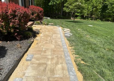 A partially completed stone walkway beside a lawn and red bushes, under bright sunlight.