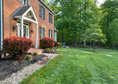 Two-story brick house with shrubs and a manicured lawn bordered by trees and a mulched garden bed.