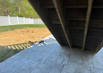 A shaded patio under wooden stairs with a grass yard and white fence in the background.