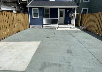 A fenced backyard with fresh concrete pavement in front of a blue house with a white door and porch.