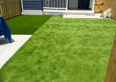 A backyard with artificial grass, a blue house, white porch, fence, and bags of supplies in the corner.