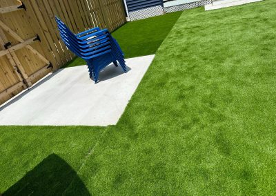 Four stacked blue chairs on a concrete slab surrounded by artificial green grass in a fenced yard.