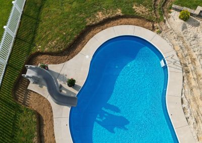 Aerial view of a kidney-shaped pool with a slide, surrounded by grass and a white fence.