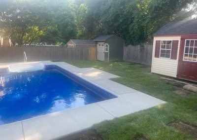 Backyard with an in-ground pool, sheds, grass, and a wooden fence surrounded by trees.