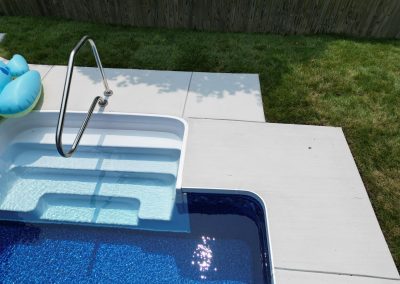 Pool steps with handrail lead into clear blue water; pool floats are on the steps, grass and patio nearby.