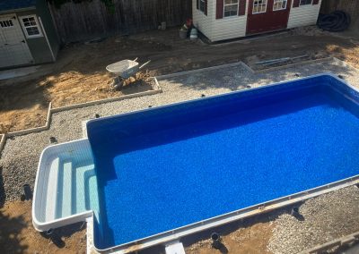 A backyard pool under construction, surrounded by gravel and dirt, with a nearby wheelbarrow and two sheds.