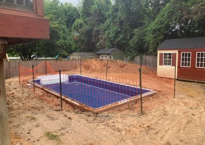 A fenced-off, in-ground pool under construction in a sandy backyard with sheds and trees in the background.