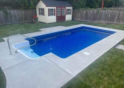 Rectangular backyard pool with concrete deck, lawn, and a small shed by a wooden fence.