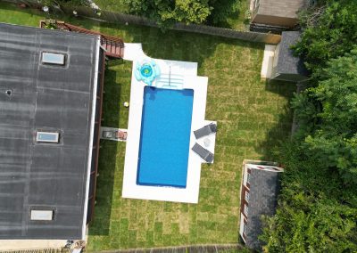 Aerial view of a backyard with a rectangular pool, pool floats, and green lawn surrounded by houses and trees.
