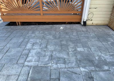 Gray stone patio with rectangular and square tiles, a wooden fence, and a small piece of trash on the ground.