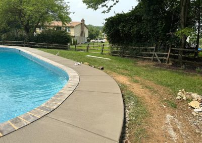Curved concrete poolside with grass, dirt path, and wooden fence, trees, and a house in the background.