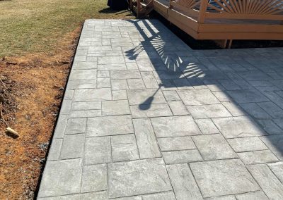Gray stone patio next to a deck with sunburst rail design, casting shadows on the patio surface.