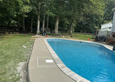 A backyard pool with scattered trash and toys on the grass and concrete near trees and a trampoline.