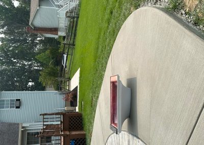 Backyard with a curved pool, concrete patio, green lawn, and a white house with a deck and fence in the background.