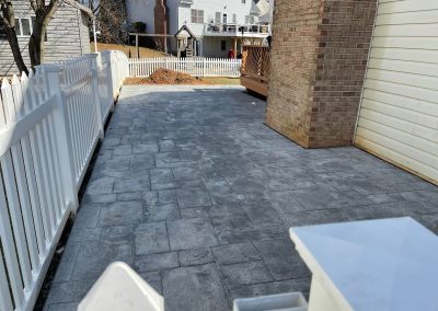 A paved patio with gray stone tiles next to a brick house and white picket fence.