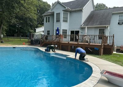 Two workers kneel by a backyard pool, repairing the edge near a house with a deck and patio furniture.