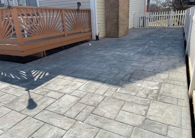 Stamped concrete patio next to a wooden deck, with a white fence and brick chimney in the background.