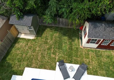 Aerial view of a backyard with two sheds, patchy grass, two lounge chairs, and part of a swimming pool.