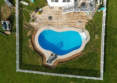 Aerial view of a fenced backyard with a kidney-shaped pool, patio, and green grass surrounding the area.