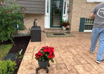 Red flowers in a pot on a patio; man in jeans walks away from a front door with plants and decorations.