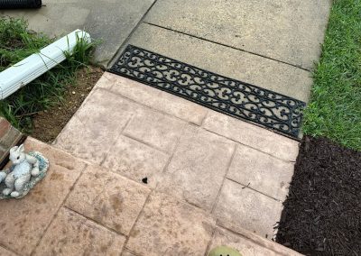 Front porch steps with a decorative black doormat, concrete walkway, grass, and a bunny statue on the left.