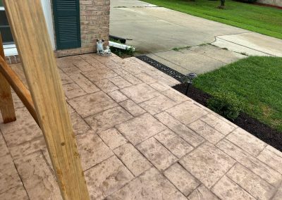 Stamped concrete patio with wooden post, small statue, and driveway next to grass and mulch.