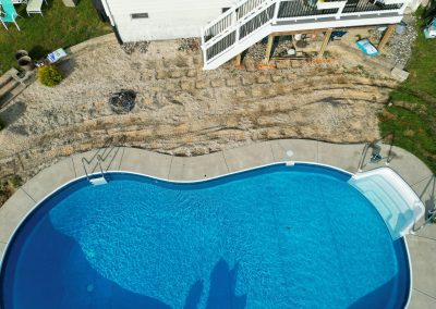 A backyard with an in-ground pool, concrete patio, and unfinished landscaping near a white house and stairs.