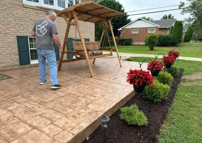 An older man walks on a stamped concrete patio with a wooden swing and red flowers along the edge.