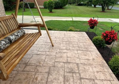 Wooden porch swing and hanging flower baskets on a stamped concrete patio overlooking a grassy yard.