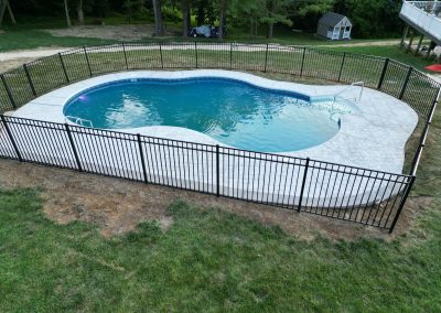 A fenced-in backyard pool with a concrete deck, surrounded by grass and trees.