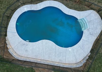 Aerial view of a kidney-shaped swimming pool with a slide, surrounded by a concrete patio and black fence.