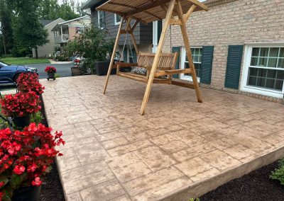 Wooden porch swing on a stamped concrete patio, bordered by red flowers in front of a house.