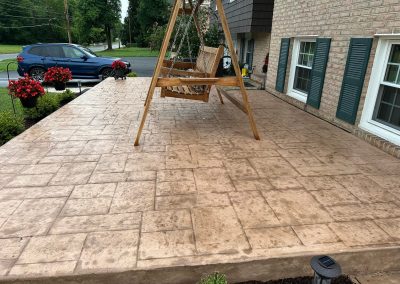 Wooden porch swing on a stamped concrete patio in front of a brick house with red potted flowers.