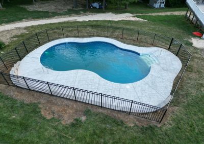A kidney-shaped backyard pool with a white deck, surrounded by a black metal fence on green grass.