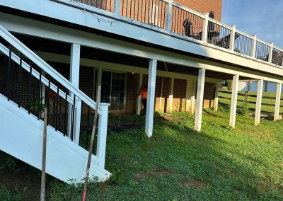 Person working under a raised white deck attached to a brick house with green grass and tools nearby.
