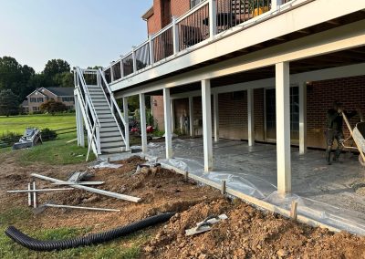 Concrete patio construction under a raised deck, with tools and materials scattered on the ground.