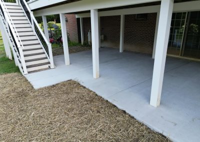Concrete patio under a deck with white columns, adjacent to straw-covered ground and staircase.