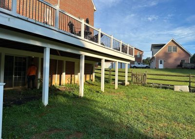 A backyard view of a house with a raised deck, grass lawn, and a wooden fence on a sunny day.