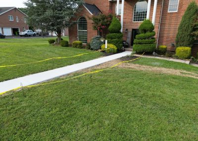 New concrete sidewalk bordered by yellow caution tape in front of a brick house with trimmed bushes.