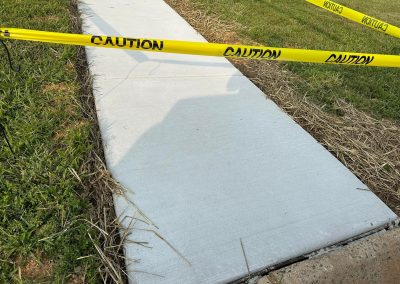 Freshly poured sidewalk with yellow caution tape around it and grass visible on both sides.