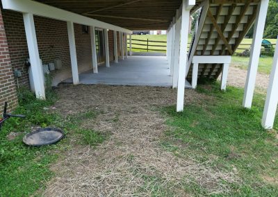Under a raised deck, a concrete patio meets a patch of straw-covered ground and green grass.