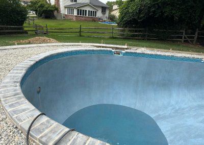 Empty in-ground pool under construction in a backyard, with grass, trees, and houses in the background.