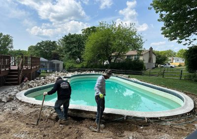 Two workers dig near an outdoor pool under a partly cloudy sky in a suburban backyard.