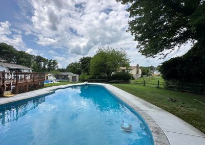 Outdoor swimming pool with clear water, surrounded by grass, trees, and a wooden fence under a cloudy sky.